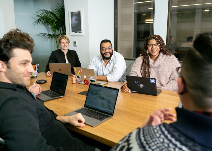 four people all on laptops, two men and two women, listen to person talking in a board meeting